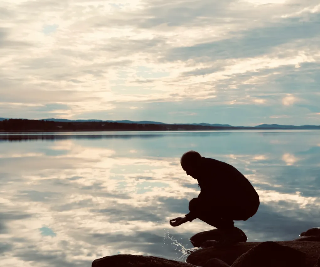 En man som sitter på huk i motljus framför en spegelblank sjö.
